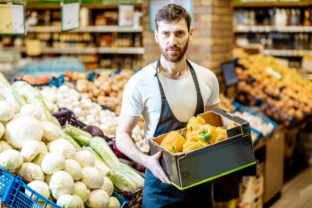 Portrait of a handsome shop worker or farmer holding box with fresh peppers in the vegetable department in the supermarket