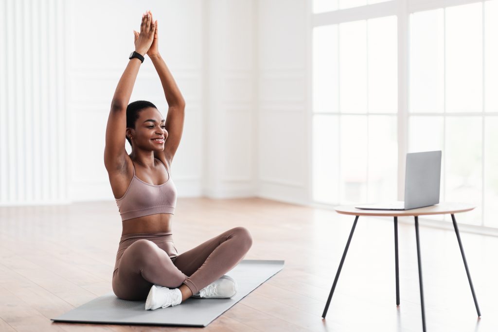 Smiling young black woman sitting on yoga mat using laptop Store App